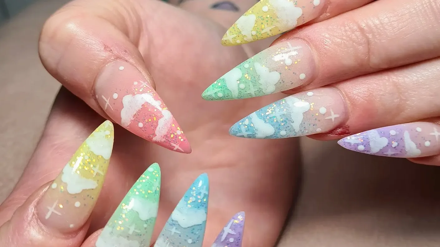Two hands of pastel rainbow cloud-and-sky stiletto nails — white clouds and glitter on a beige ground
