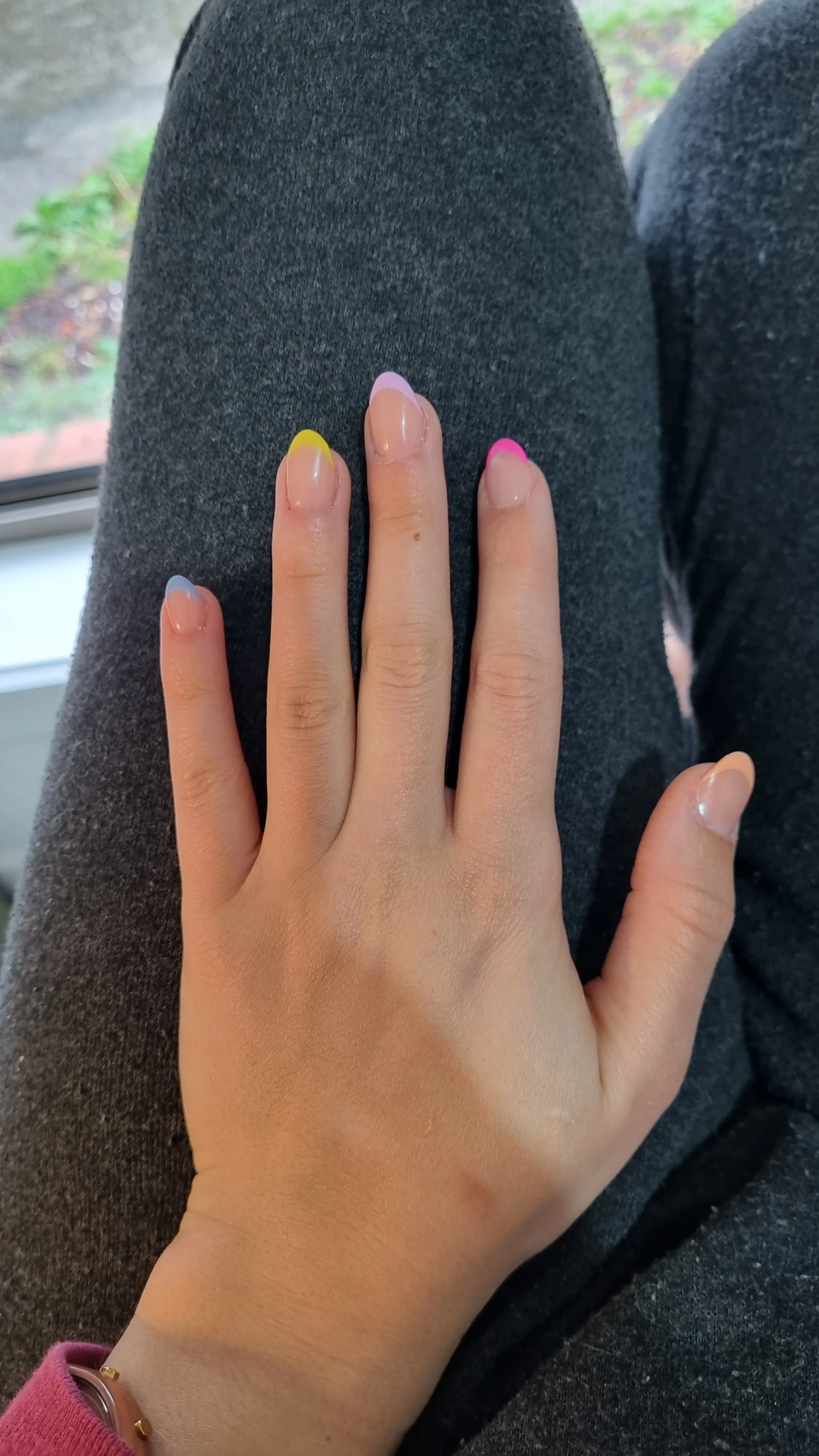Pastel coloured French tips — yellow, lilac, pink, blue — on almond nails, hand on grey knit