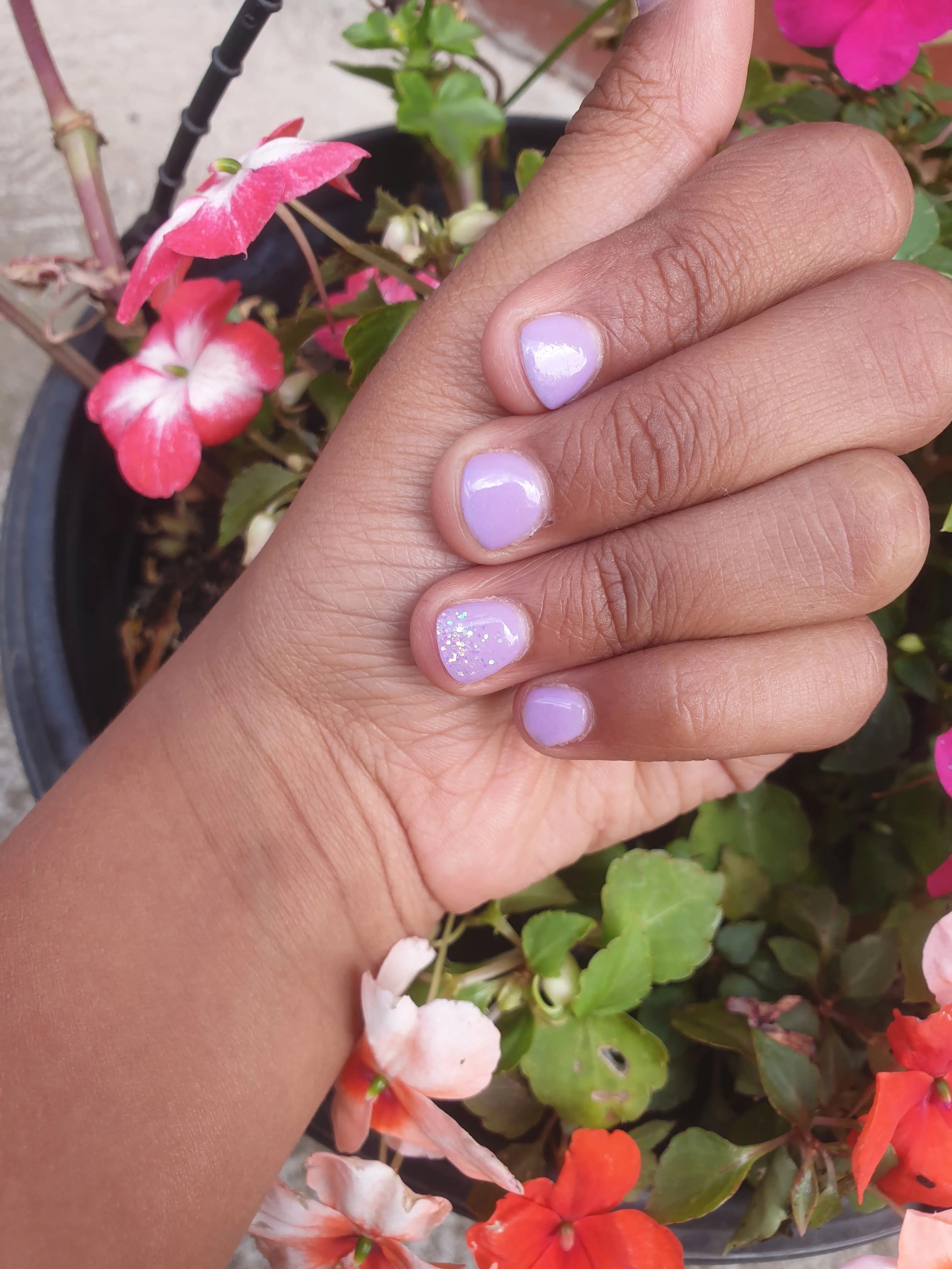 Lilac nails with an iridescent-glitter accent nail, hand held against potted impatiens with pink and coral blooms