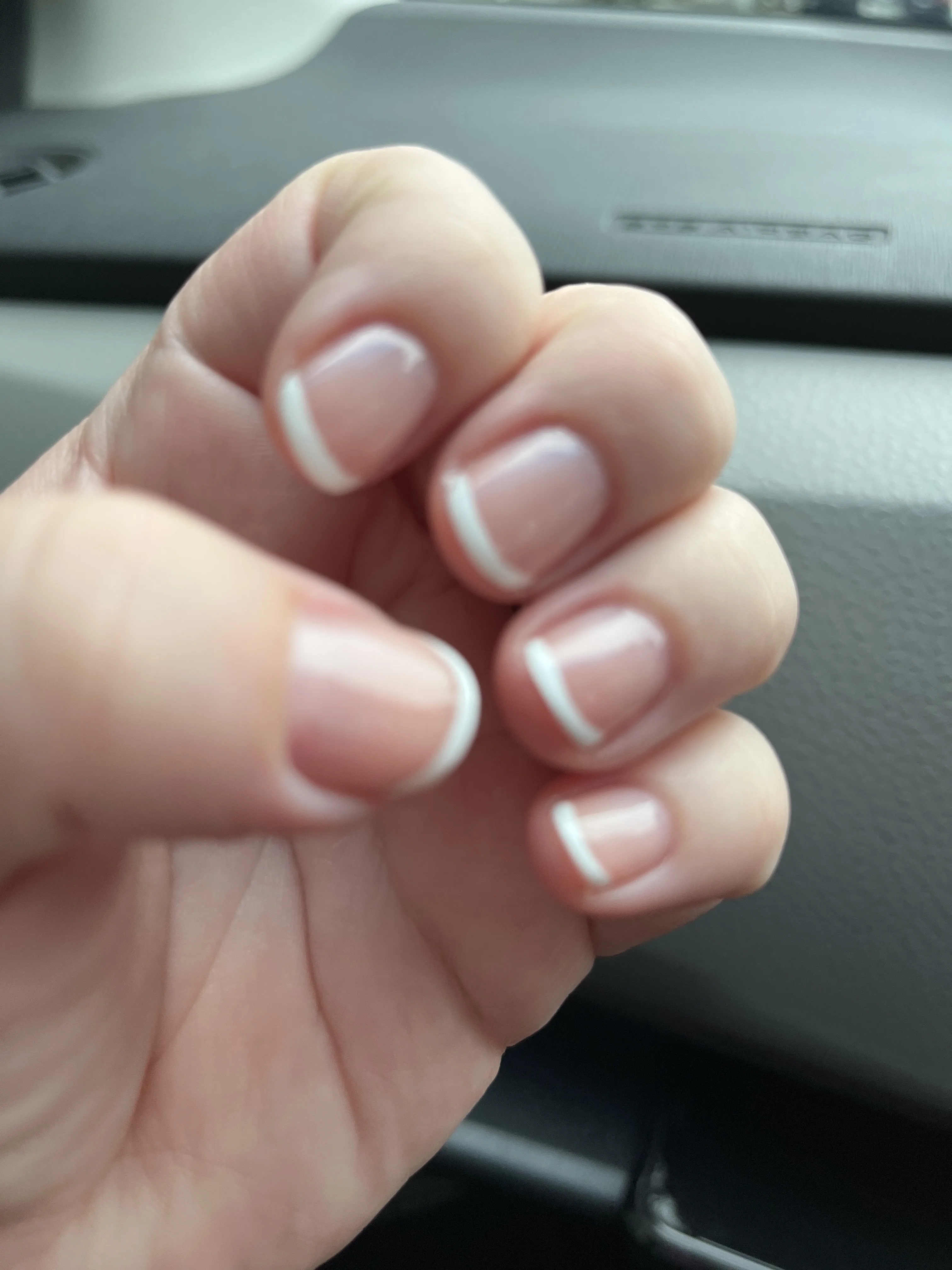 Natural pink nails with slim white French tips, classic set photographed with a car dashboard behind