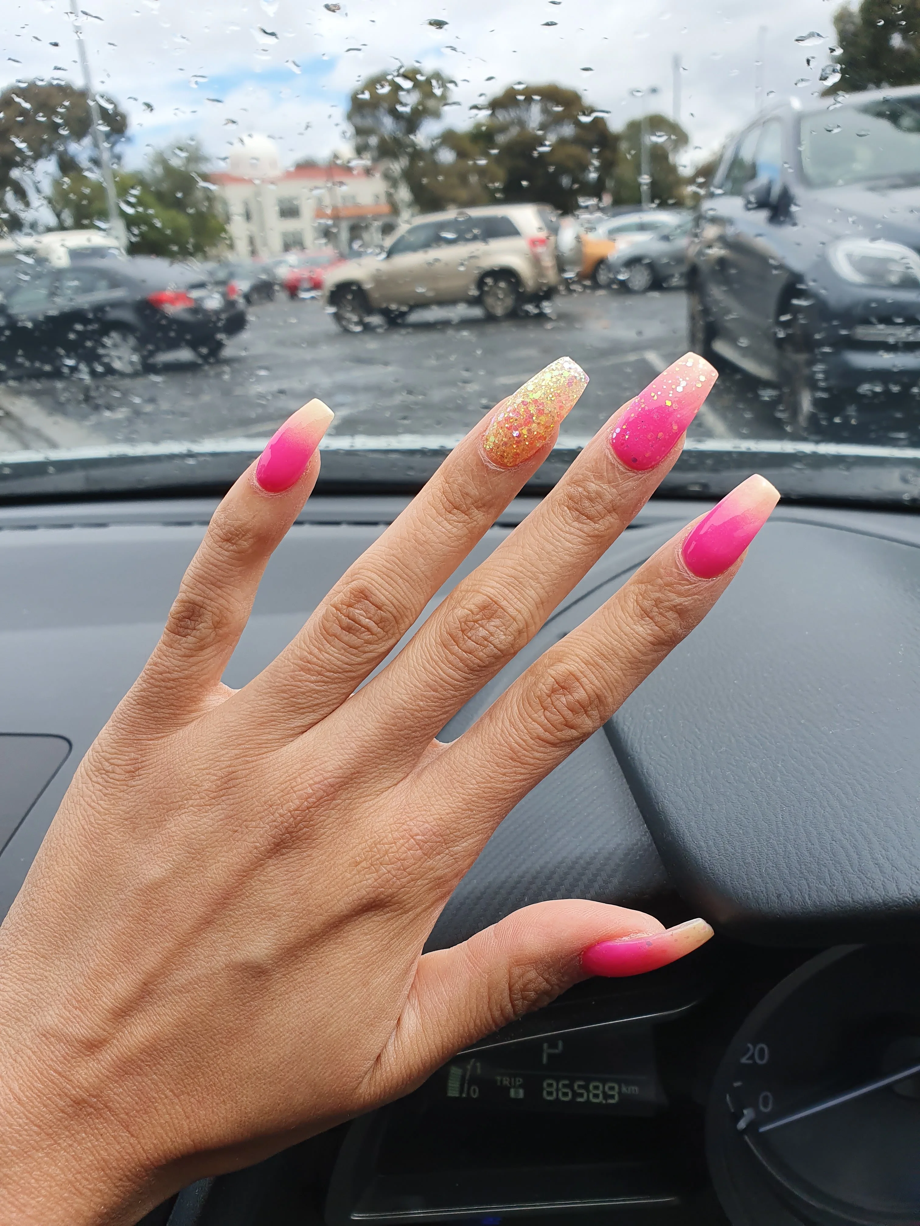 Pink and gold-glitter coffin nails photographed from a car, with a rain-flecked windscreen over a Coburg carpark and a white-domed building behind
