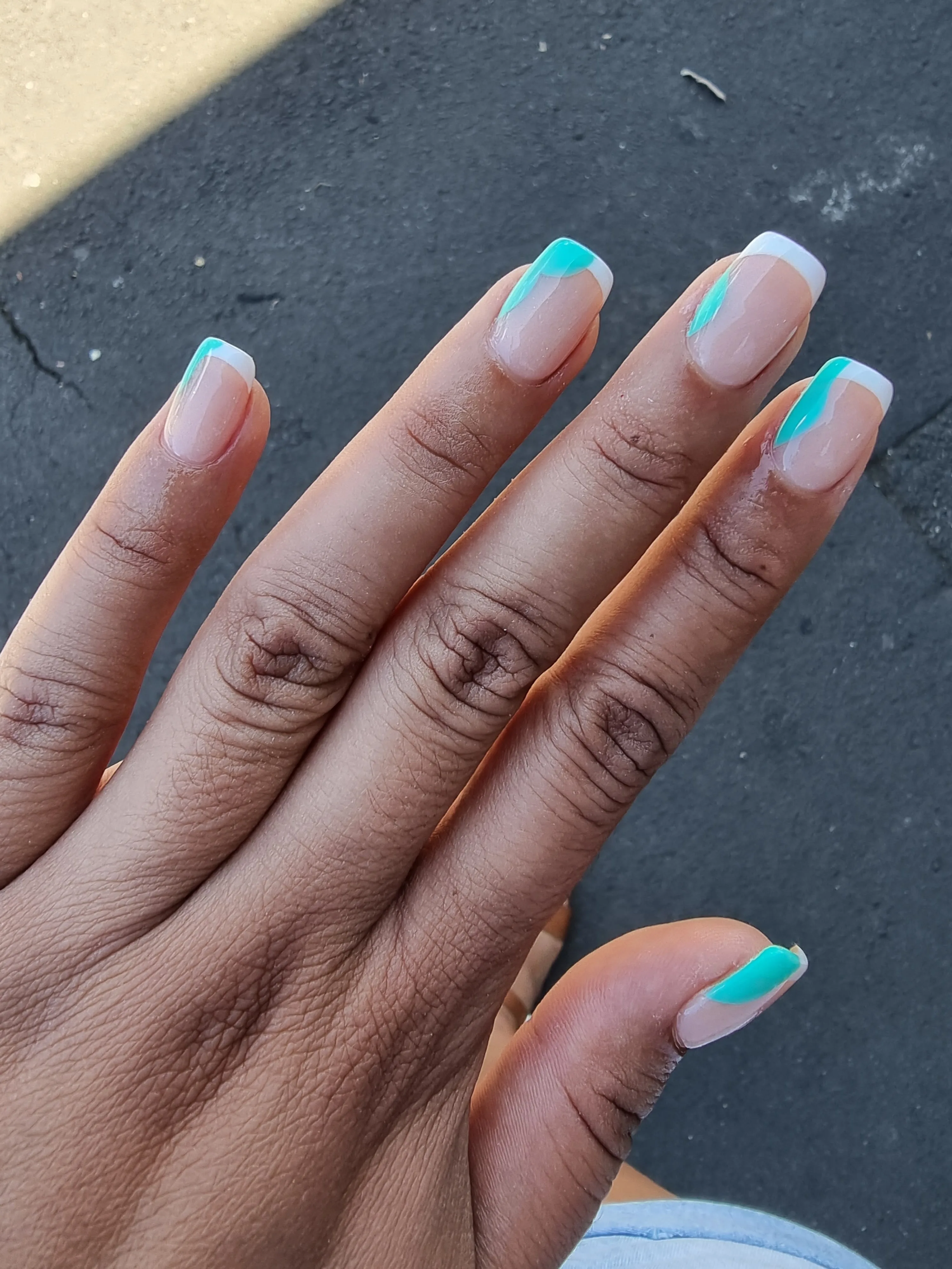 Teal-swirl French tips on a deep-skin hand with outdoor asphalt behind