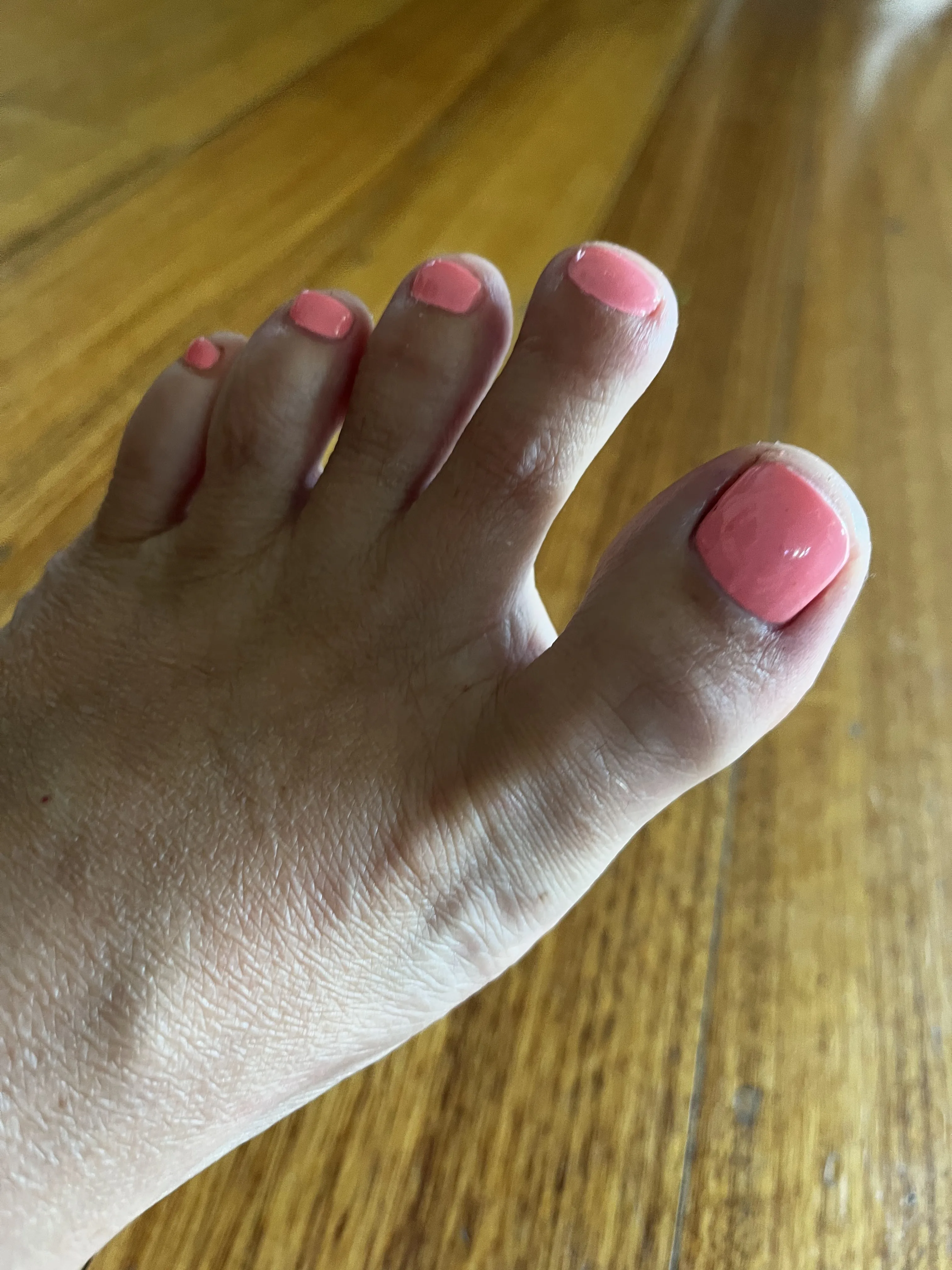 A foot with square coral-polished gel toenails on a timber floor — pedicure finish