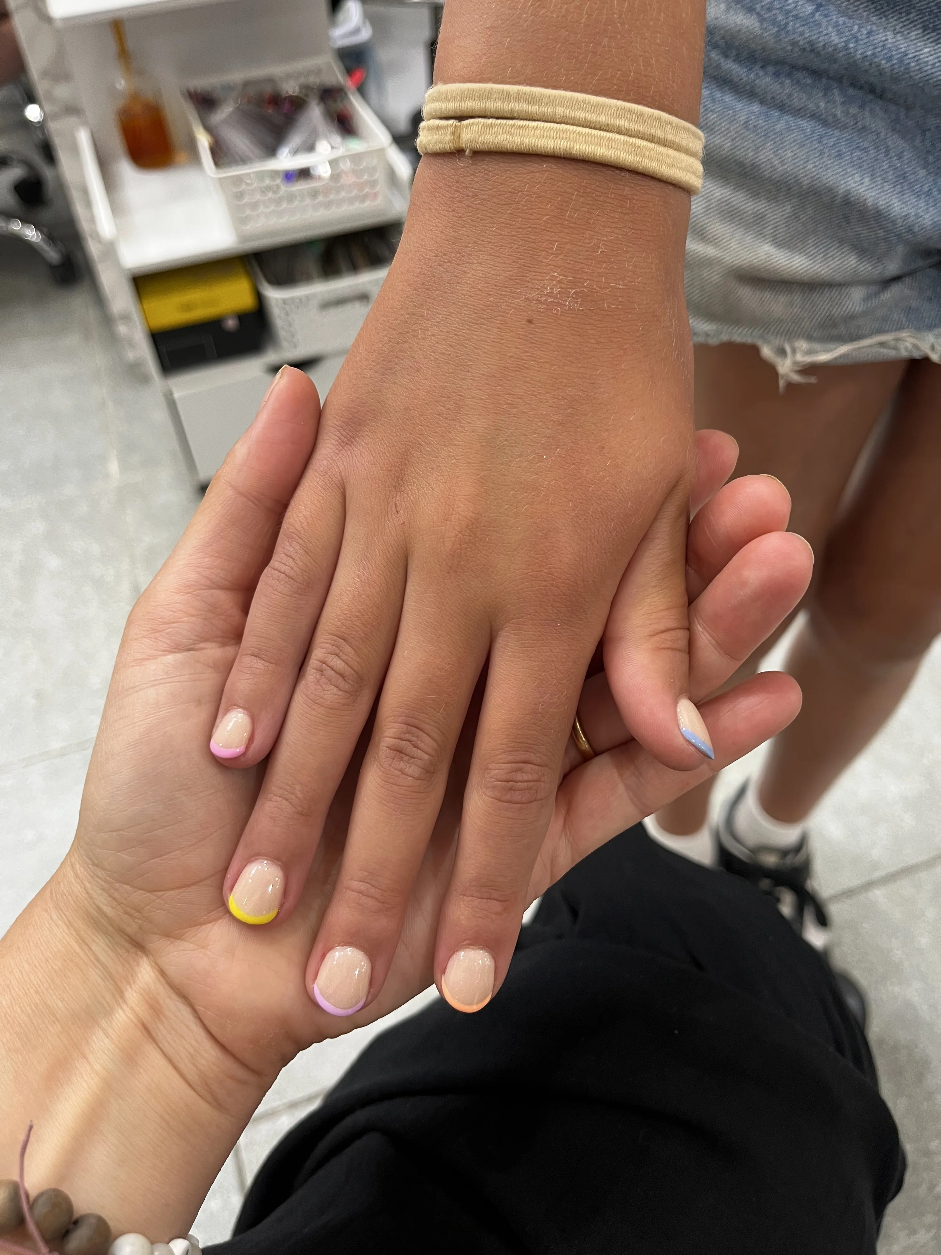 Two hands showing pastel micro-French coloured tips in a soft salon setting with a polish rack in soft focus