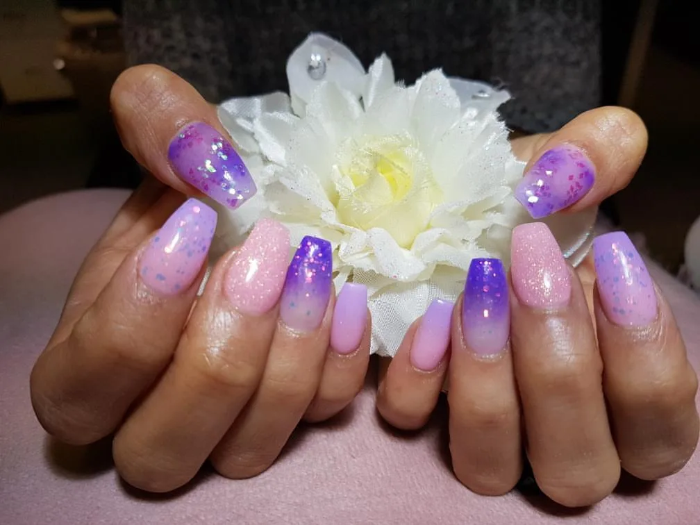 Cupped hands in lilac and pink glitter ombre square nails around a white silk flower on a pink cushion